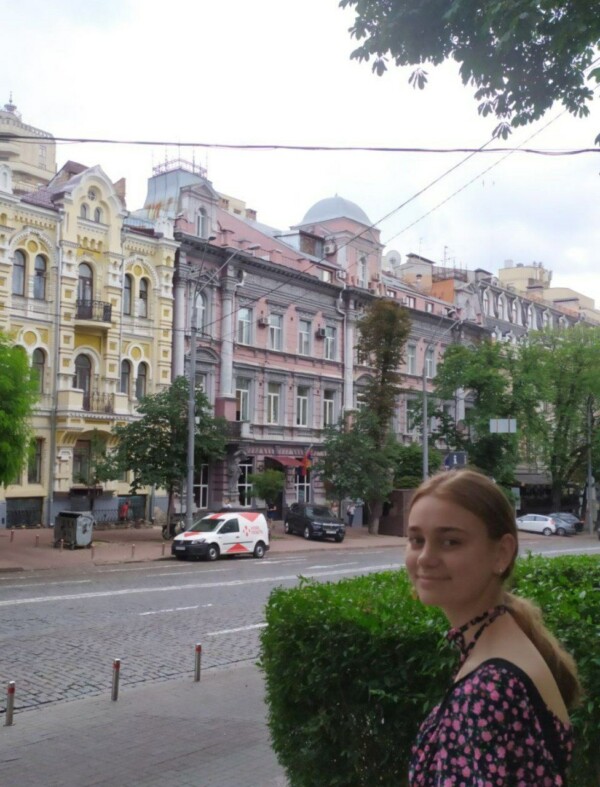 Daria, smiling, is standing in front of a Street in Kharkiv. In the background you can see the road, parking cars and multistory buildings in different colors