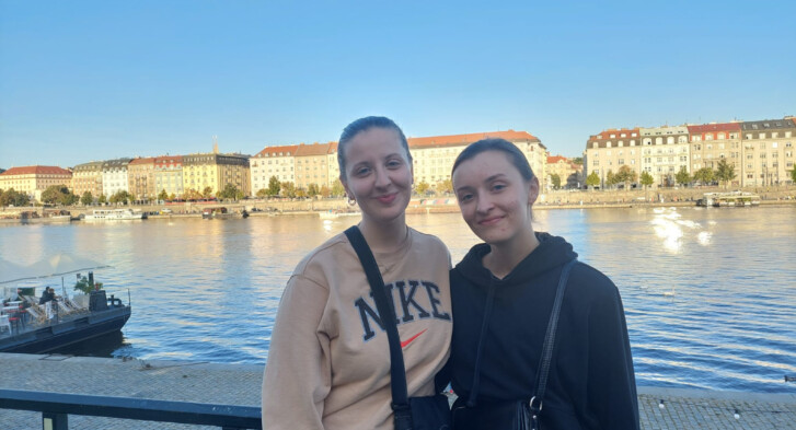 Two women standing near the Vltava river, smiling.